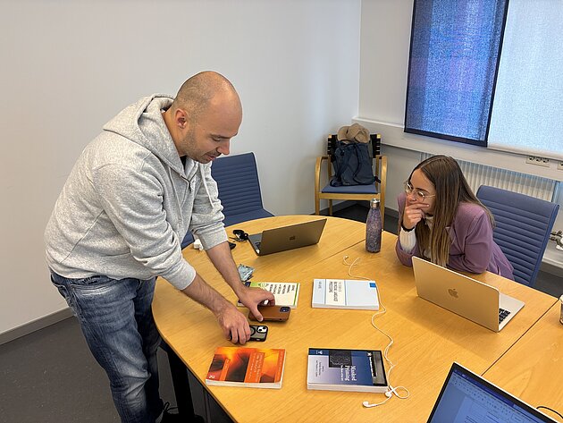 Two people on opposite sides of a table. They are looking at objects on the table: two phones and four books.