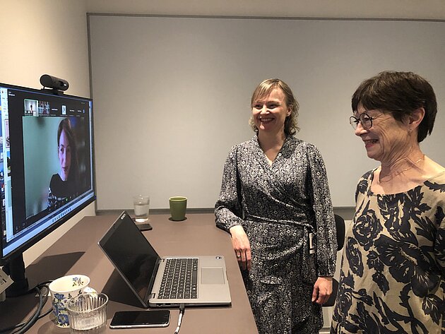 Emily Grenner and Kristina Hansson stand in front of a computer screen. Ifigenia Dosa can be seen on the screen. All three are smiling.