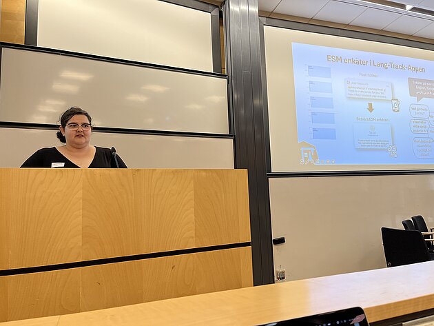 Henriette Arndt stands at the pulpit looking out at the audience. In the background you can see a PowerPoint slide with a graphic flow chart on it.