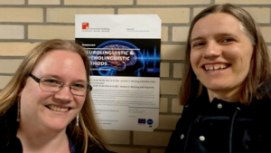 Sabine and a German colleague are looking into the camera and smiling. In the background, a poster for the training event.