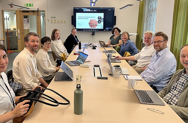 People seated around a long table. At the end of the table a big screen with a PowerPoint presentation and some people participating over zoom.