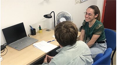 Two people seated at a table getting ready for an experiment. The participant is seen from behind, the researcher, Angelica Zordan, is looking at the participant and smiling.