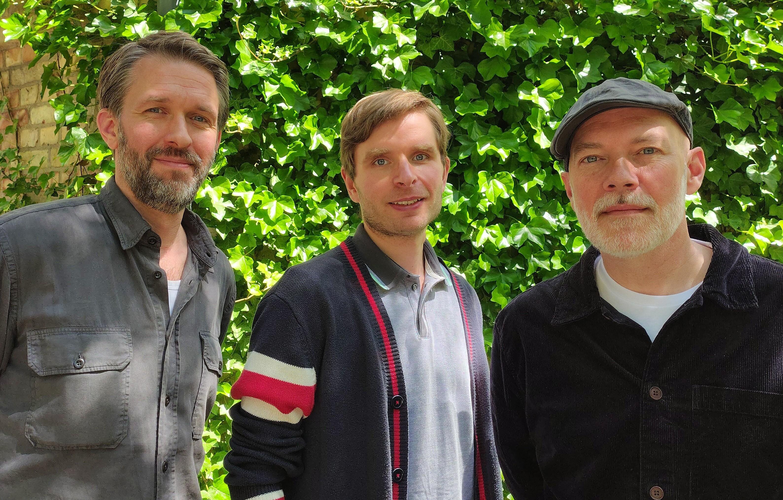 The three researchers stand outside in front of a leafy background. They are all looking into the camera.