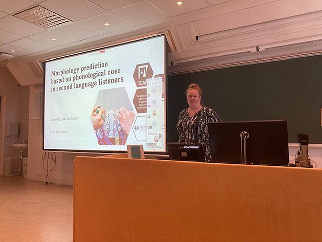 Sabine stands next to a screen showing a powerpoint presentation.