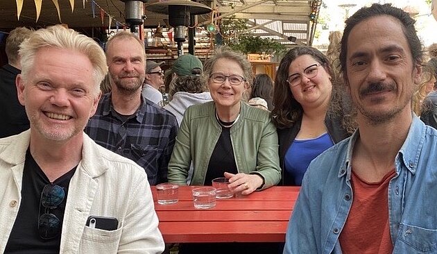 All five project members are seated around a table in a restaurant patio. They are all looking into the camera and smiling. 