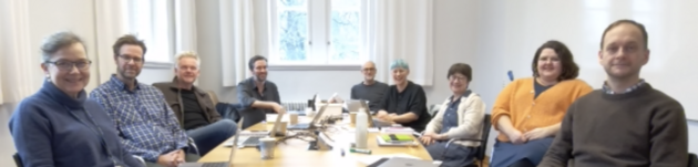 A blurry photo of everyone participating in the retreat. All nine people are seated around a table. They are smiling at the camera. The photo is blurry.